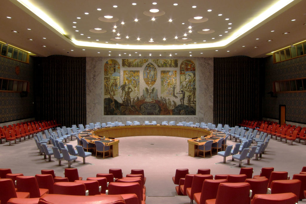 Photo of the UN Security Council chamber with a horseshoe shaped meeting table surrounded by empty chairs and a large mural painting behind the table