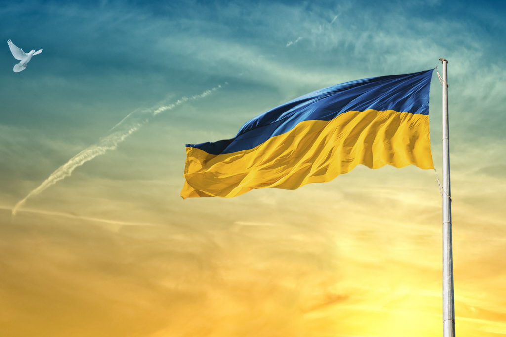 Ukraine flag (blue and yellow stripes) flying on a flag pole with a dove flying toward the flag at sunset