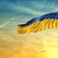 Ukraine flag (blue and yellow stripes) flying on a flag pole with a dove flying toward the flag at sunset