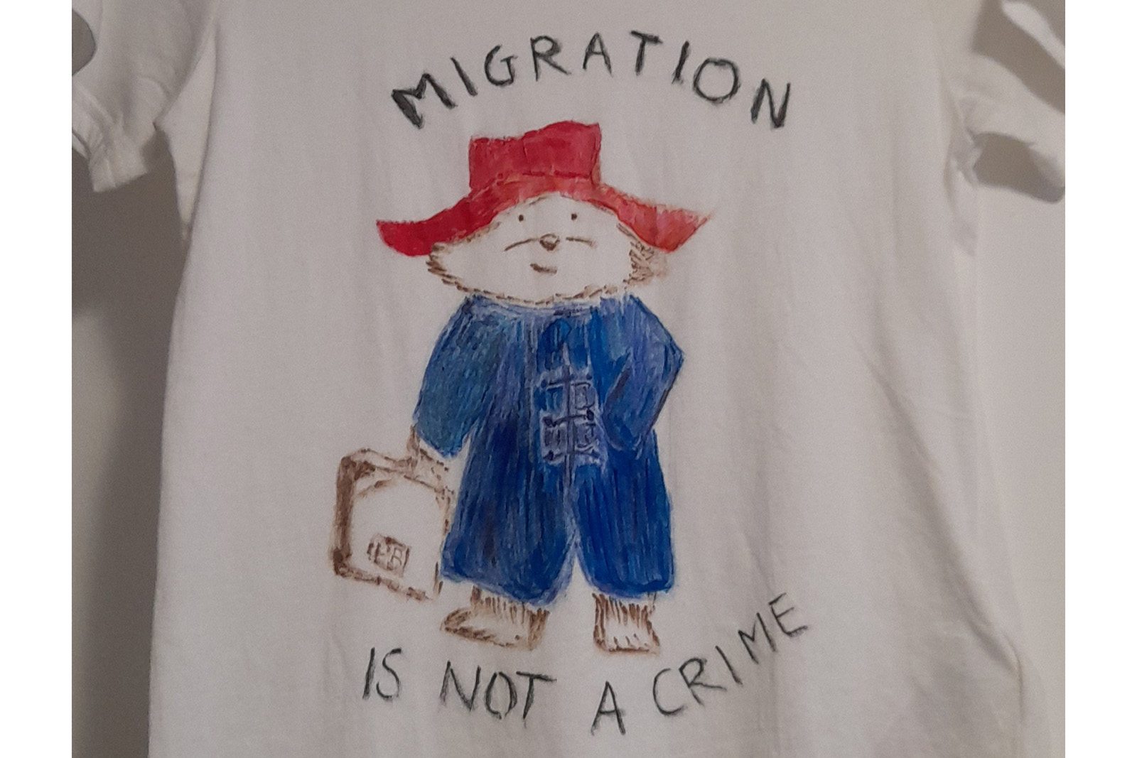 Drawing of a bear in a red hat and blue overalls carrying a suitcase on a t-shirt with "migration is not a crime" surrounding the picture