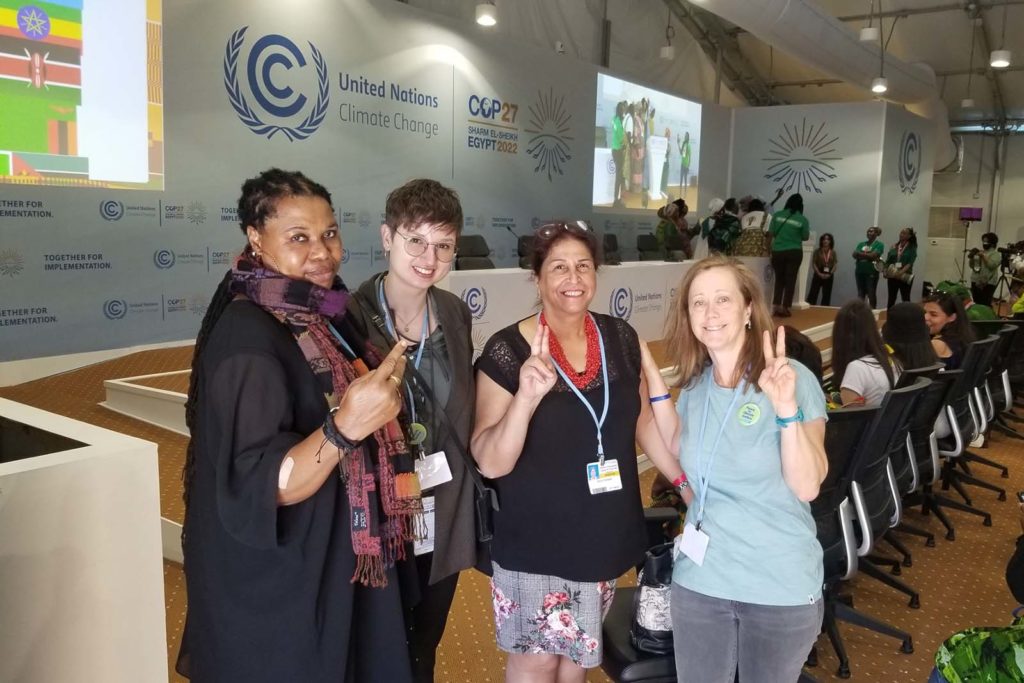 Four women making the peace sign with their fingers standing in front of a panel table with the United Nations Climate Change and COP27 logos on the wall behind the table