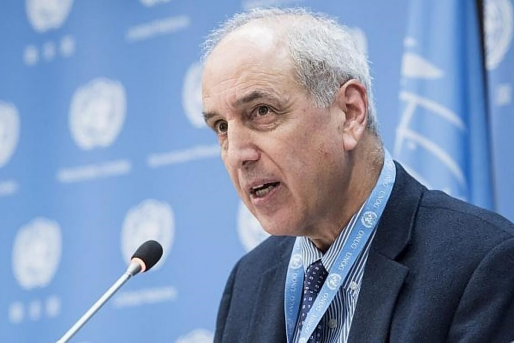 Michael Lynk speaking into a microphone with a light blue background with the UN logo.