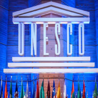 UNESCO logo that has the term UNESCO with three lines below and a roof above the name. The logo is hung in front of a blue background with a row of flags from different countries below.