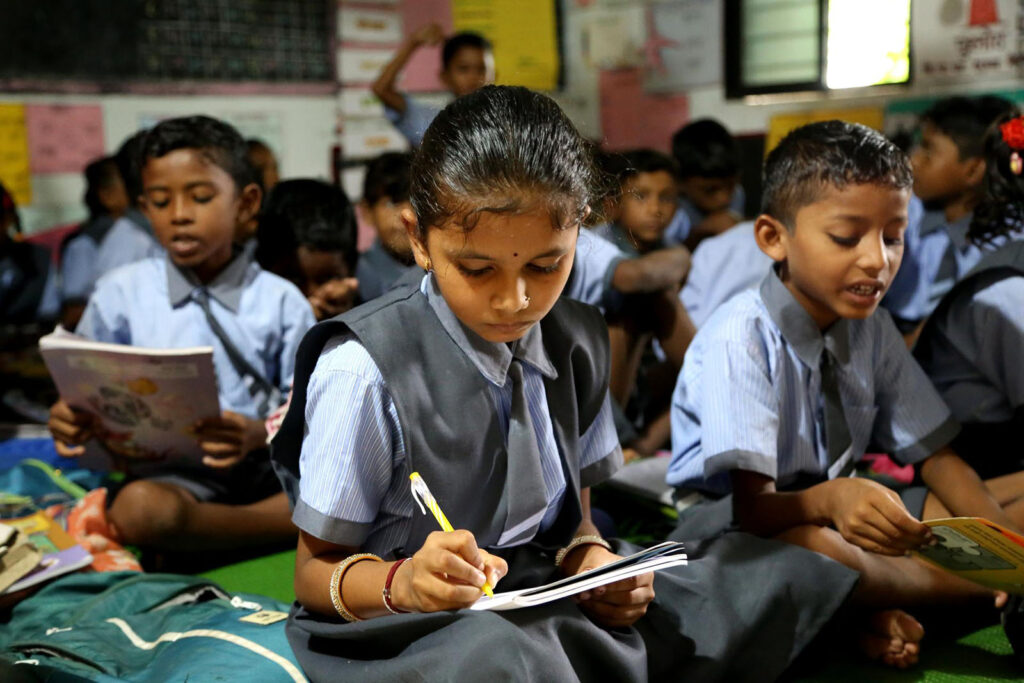 Children in school uniforms sitting on the floor reading or writing in books.