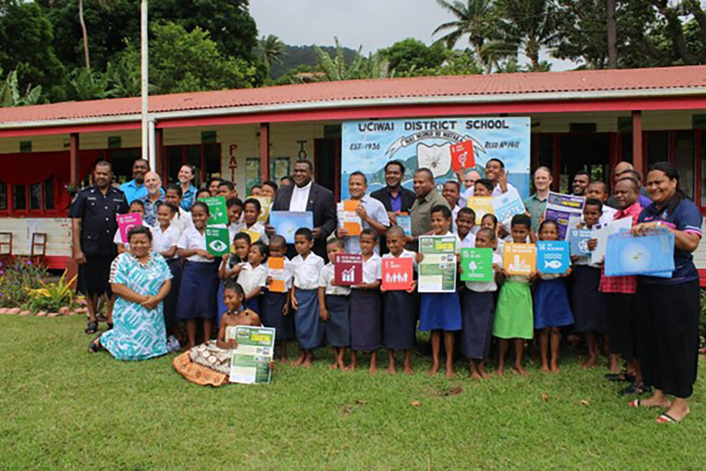 A crowd of adults and children standing in front of teh UCIWAI District School building holding signs showing the UN Sustainable Goals