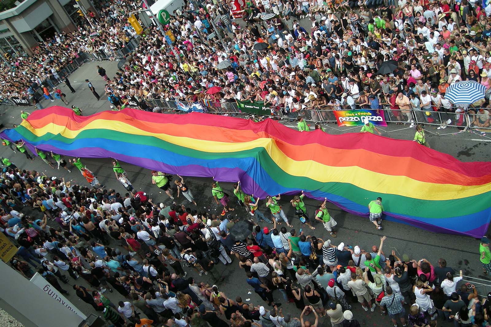 Photo looking down on a street with a long Pride flag being held by about 20 people. A crowd of people surround the flag on both sides of the street.