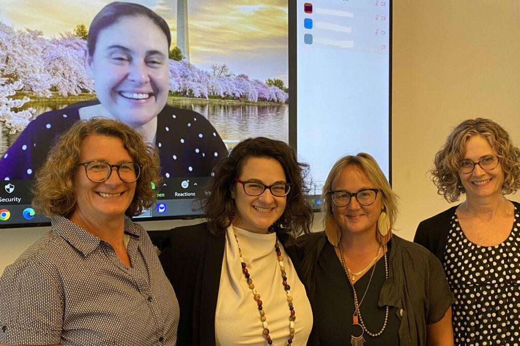 Margaret Walton-Roberts, Allison Petrozziello, Jenna Hennebry and Andrea Brown standing in front of a TV screen with Rosa Celorio. Alison Mountz is not pictured.
