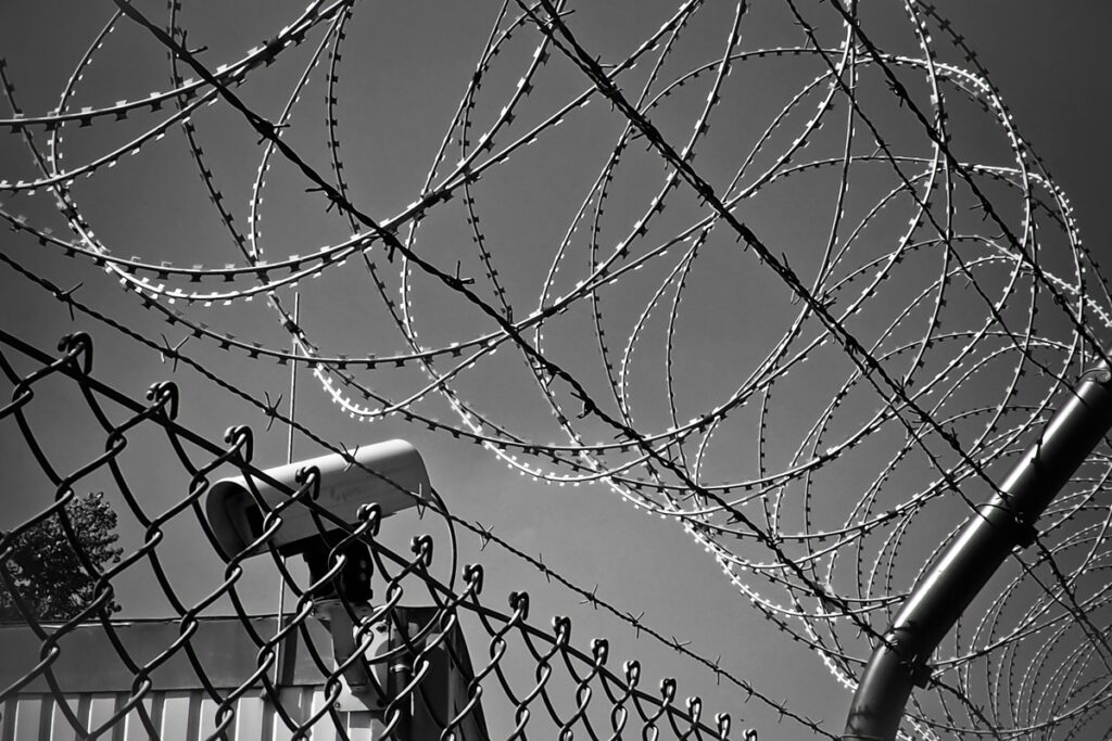 Black and white image of barbed wire over a chain link fence with a camera and top corner of a building behind the fence.