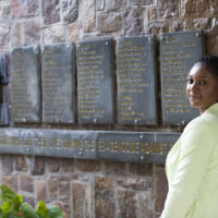 Adwoa standing next to a wall with plaques displaying names and the words, "never again"