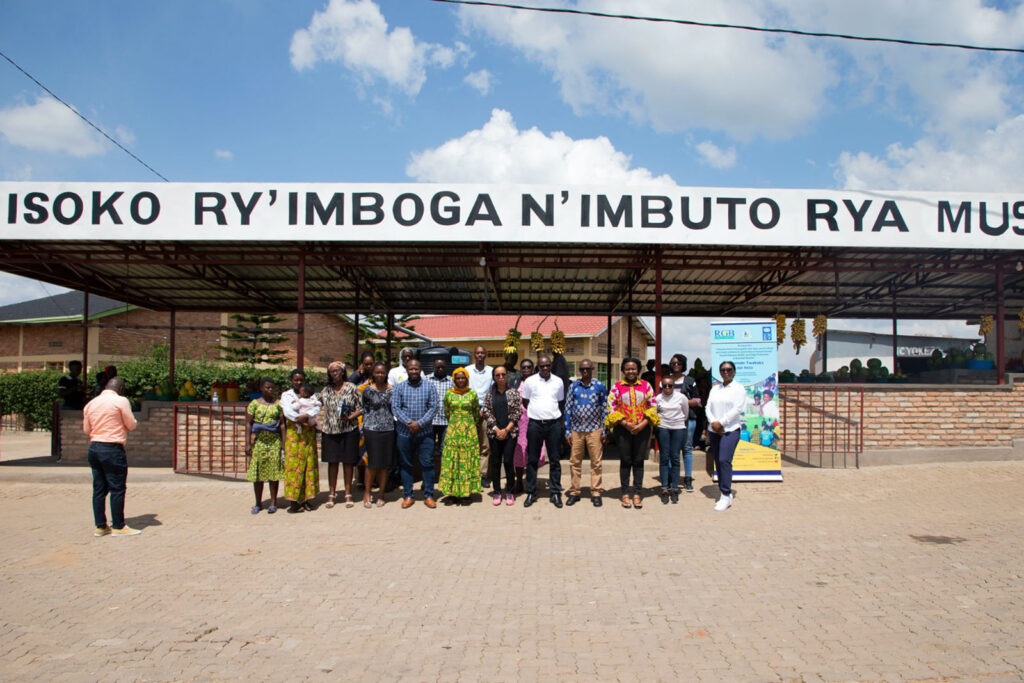 Group of people standing in front of a sing "Isoko Ry'Imboga N'Imbuto Rya..."